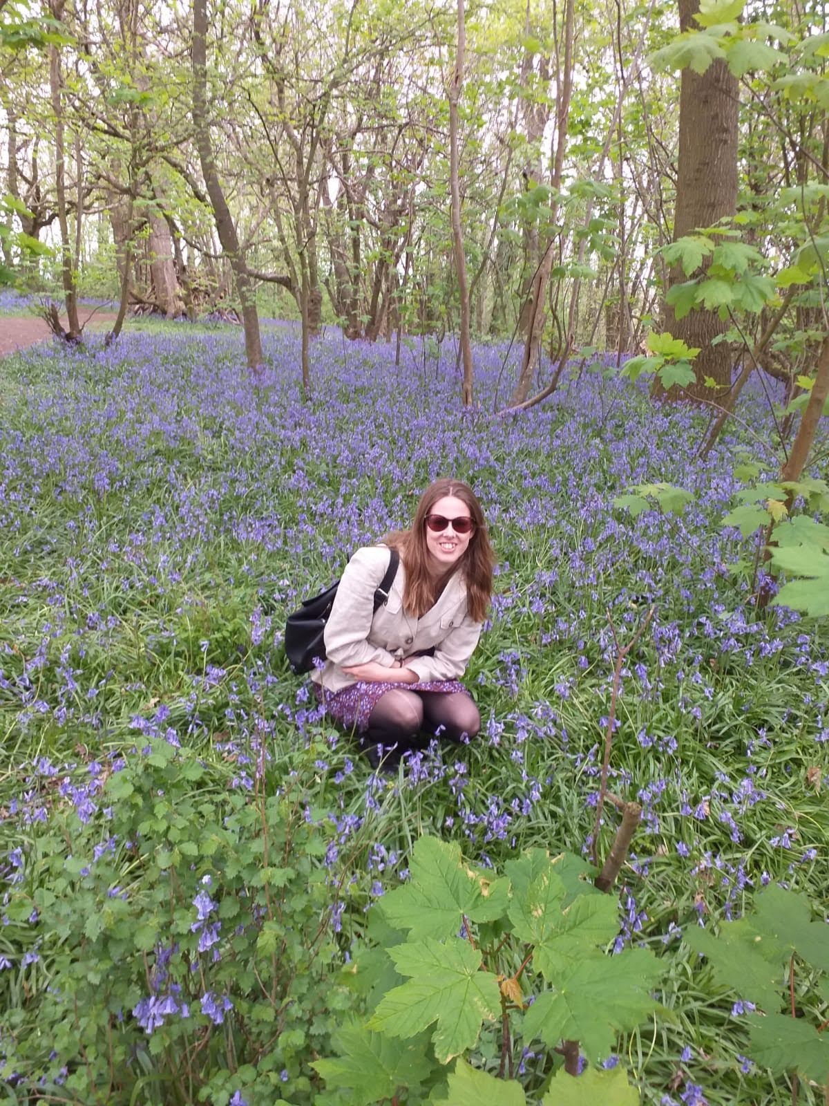Cadence, a white woman with medium length brown hair and sunglasses, wearing a beige linen jacket with a brown and purple floral skirt, and carrying a black backpack, crouches in the middle of a carpet of bluebells)